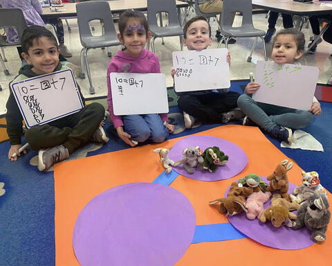 Kindergarten students pose proudly on classroom carpet holding up whiteboards with math equations