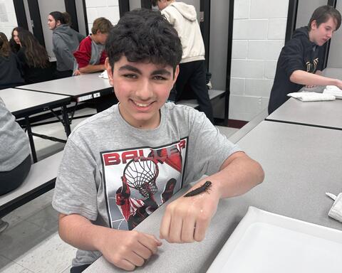 Student poses proudly holding a bug on the top of his left table while sitting at a table