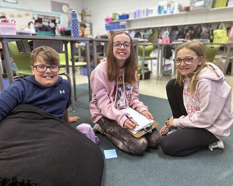 Three students smile on carpet in classroom while writing poems