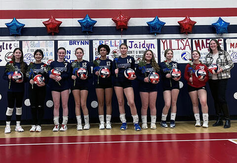 Eighth grade girls' volleyball team proudly pose with flowers, volleyballs and coach in school gym