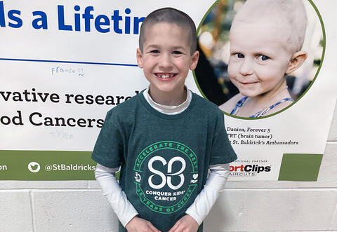 Student poses proudly against St. Baldrick's wall poster after getting head shaved at school event